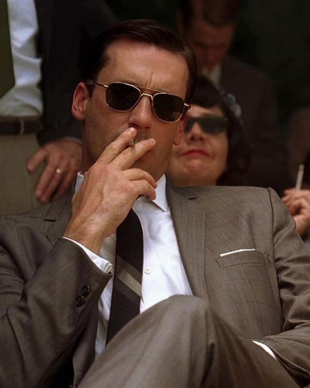 A man in a grey mid-century suit and sunglasses smoking a cigarette outdoors.