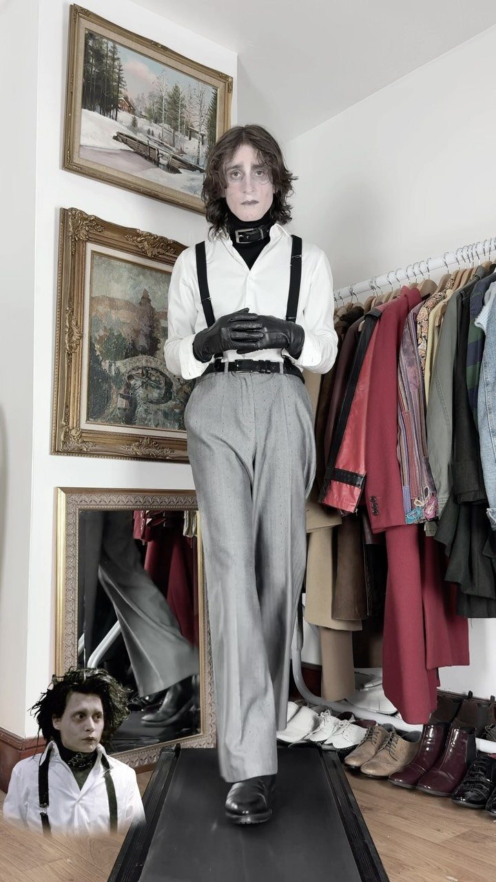 A man with pale makeup wearing a white shirt, black suspenders, and grey trousers stands indoors.