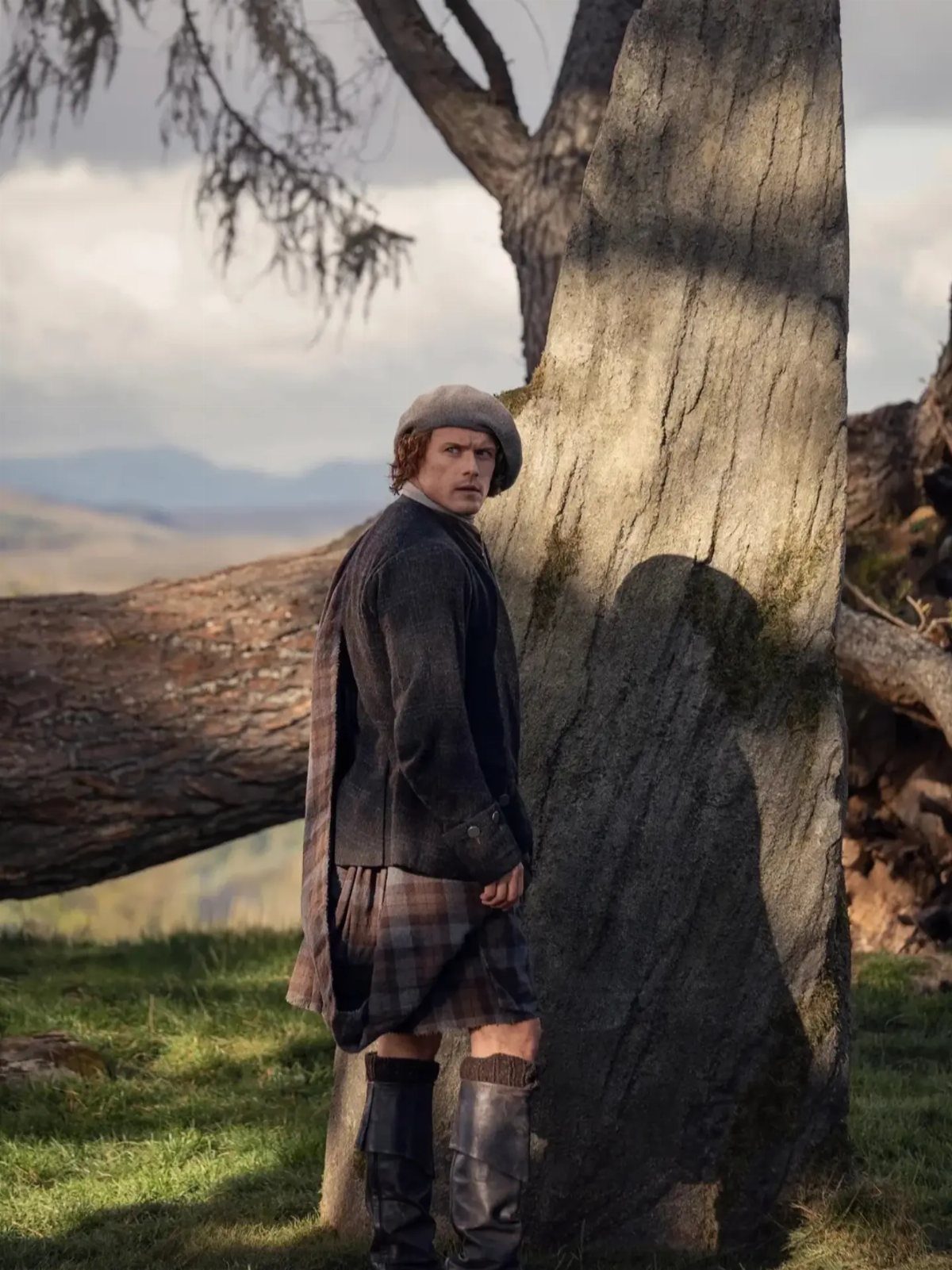 A man in historical Scottish attire including a kilt and cap stands beside a large tree.