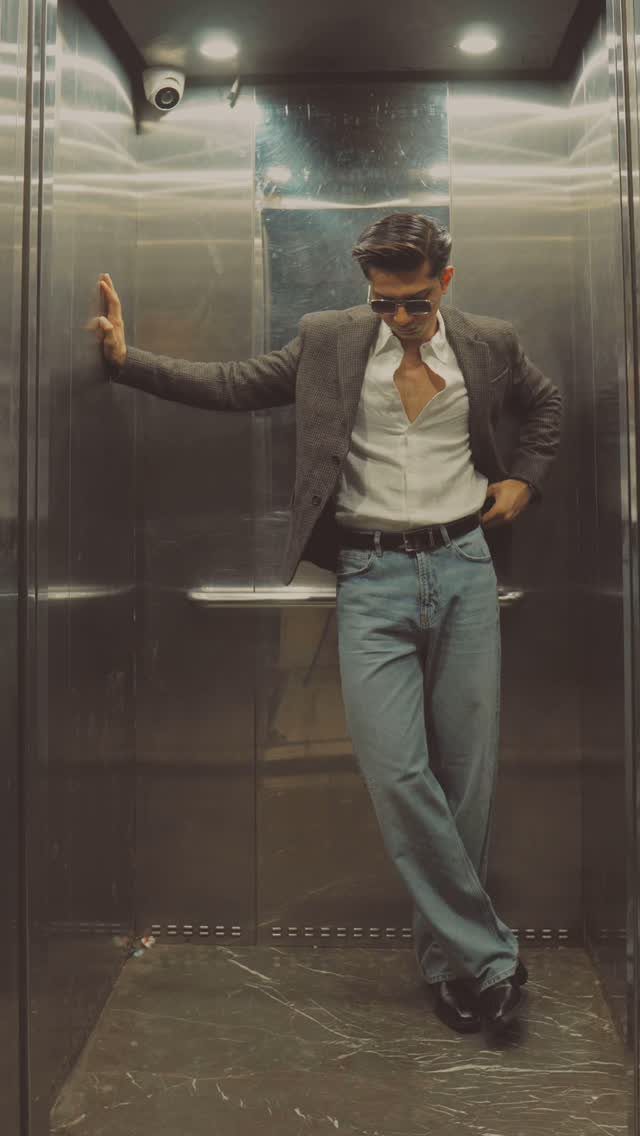 A man in a blazer, white shirt, and jeans posing inside a metallic elevator.