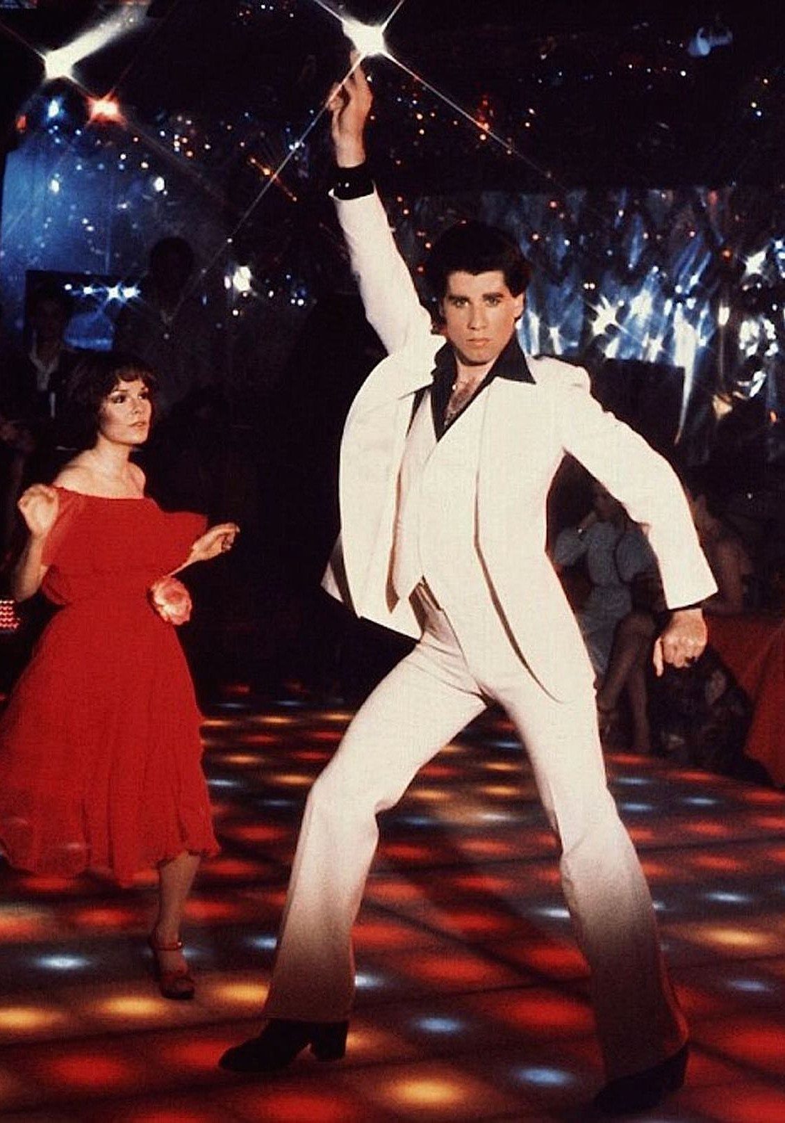 A man in a white disco suit dancing on a colorful lit dance floor.