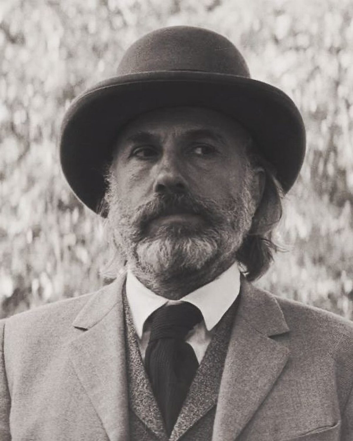 A bearded man wearing a vintage hat, suit, and tie in a black and white portrait.