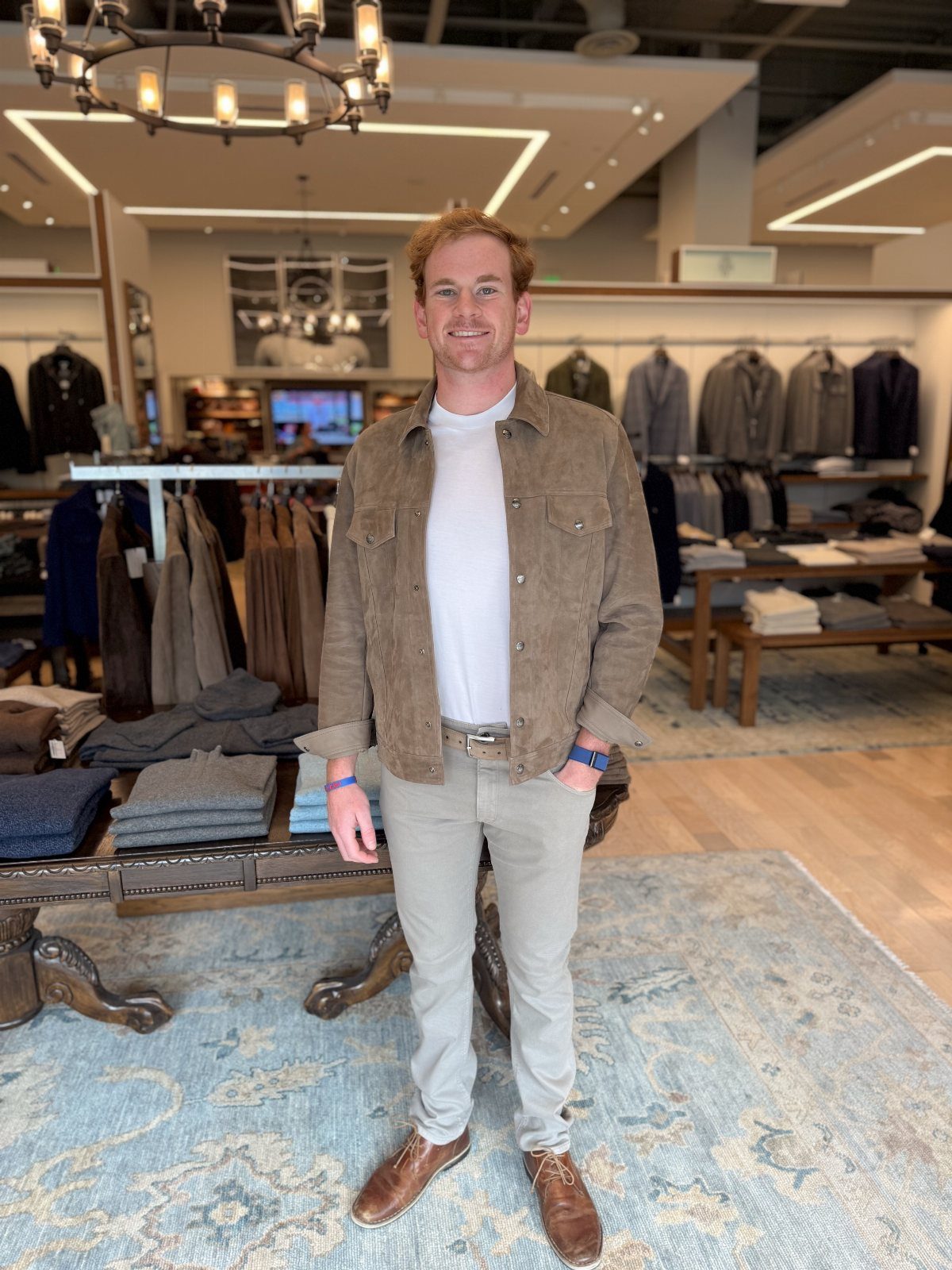 A smiling man wearing a brown suede jacket, white t-shirt, and beige trousers in a store.
