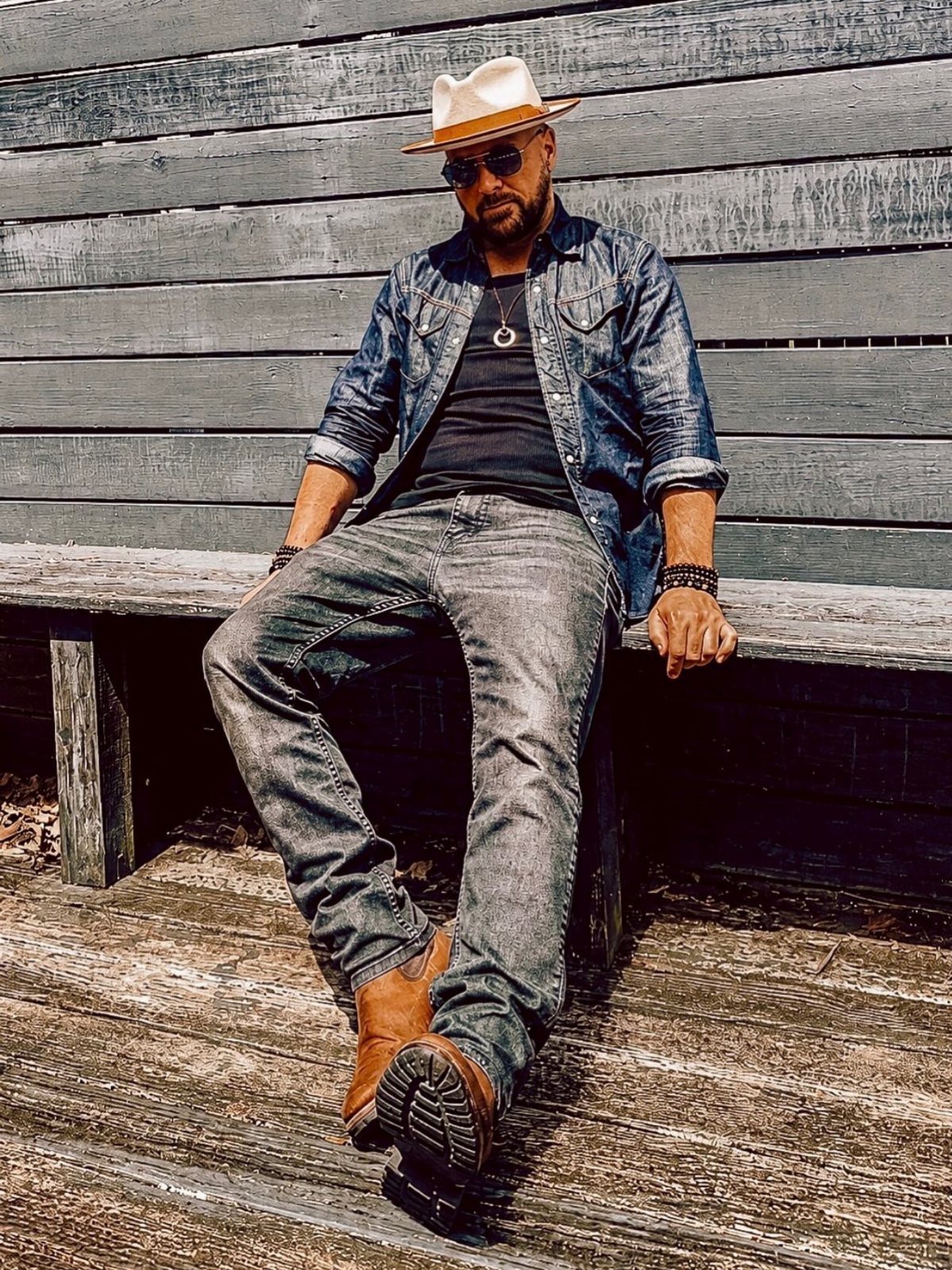 A man in a denim shirt and grey jeans sitting on a wooden bench outdoors.