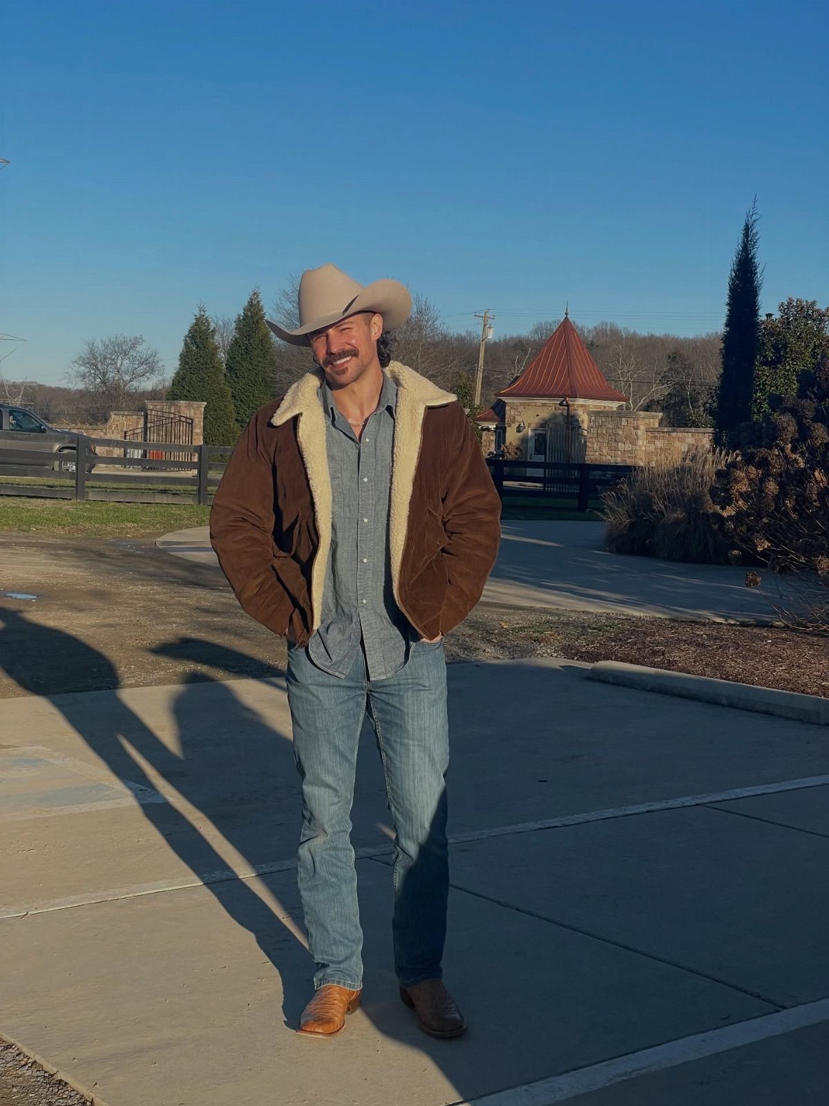 A man wearing a cowboy hat, shearling jacket, and jeans standing outdoors in sunlight.