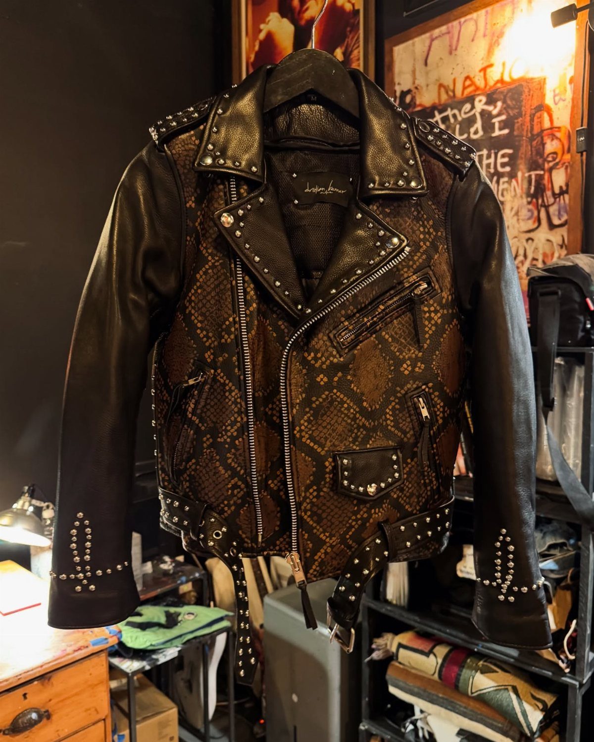 A black and snakeskin patterned leather biker jacket with silver studs hanging on a hanger.
