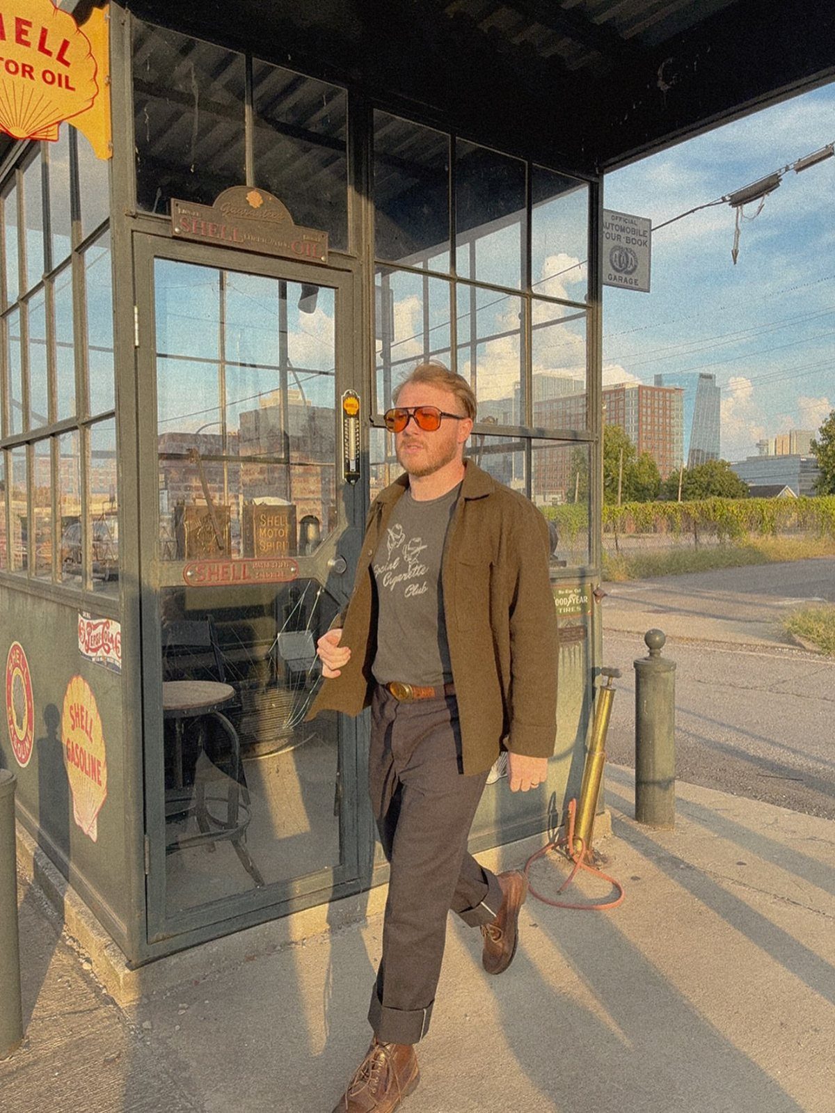 Man in sunglasses and a brown jacket walking past a vintage glass building at sunset.