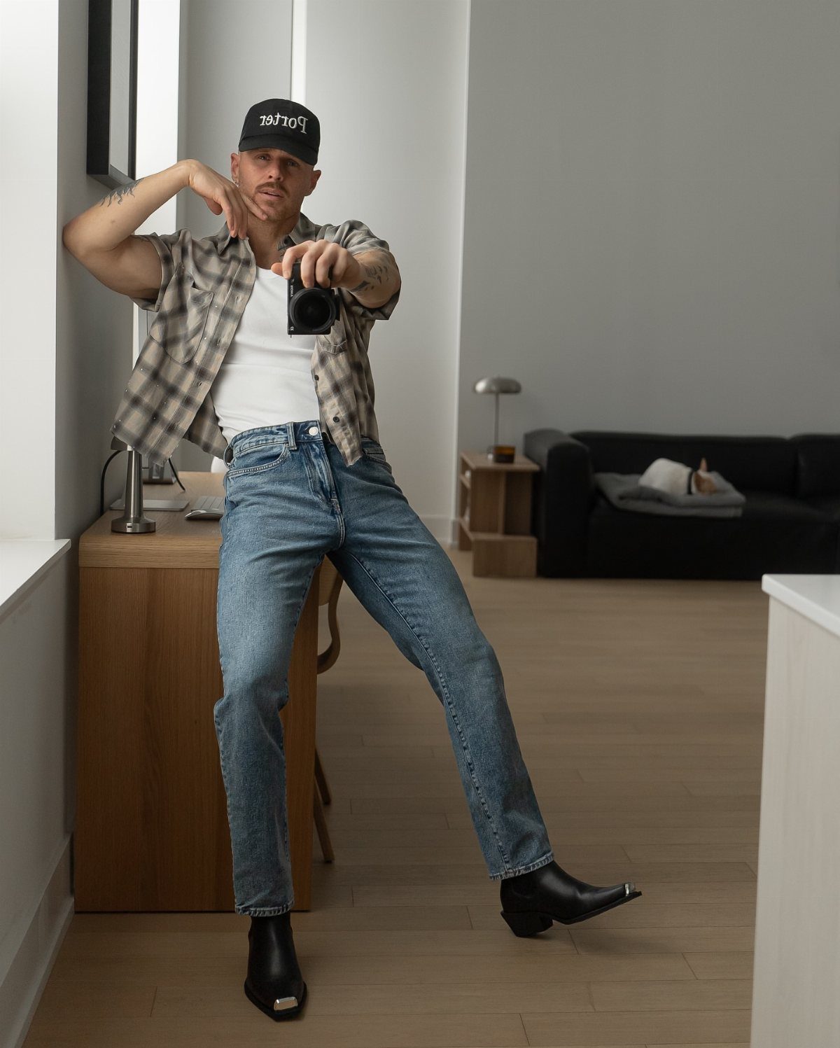 Man in plaid shirt, blue jeans, and black boots posing for a mirror selfie indoors.