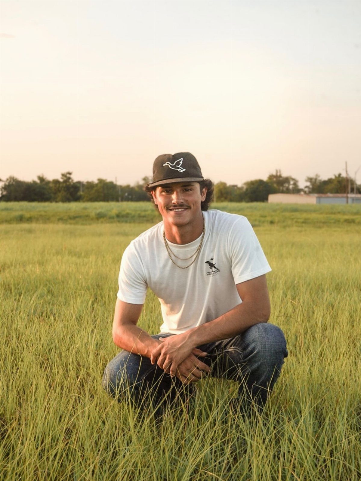 A man in a white t-shirt and jeans squats in a grassy field at sunset.