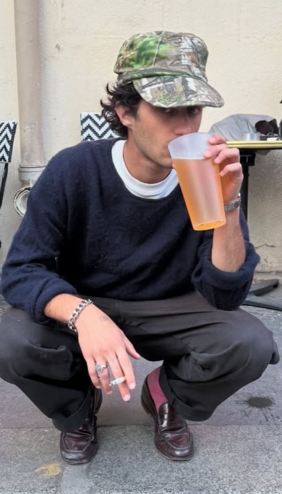 A man wearing a camouflage baseball cap, navy sweater, and dark loafers crouches outdoors.