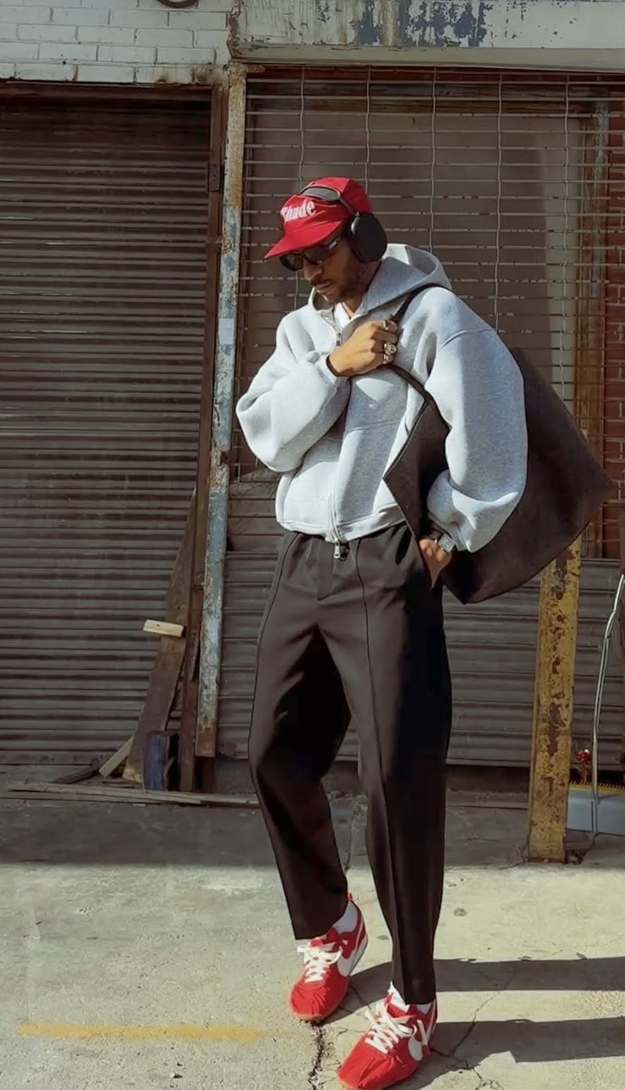 Man in grey hoodie, black pants, red cap, and red sneakers standing outdoors.