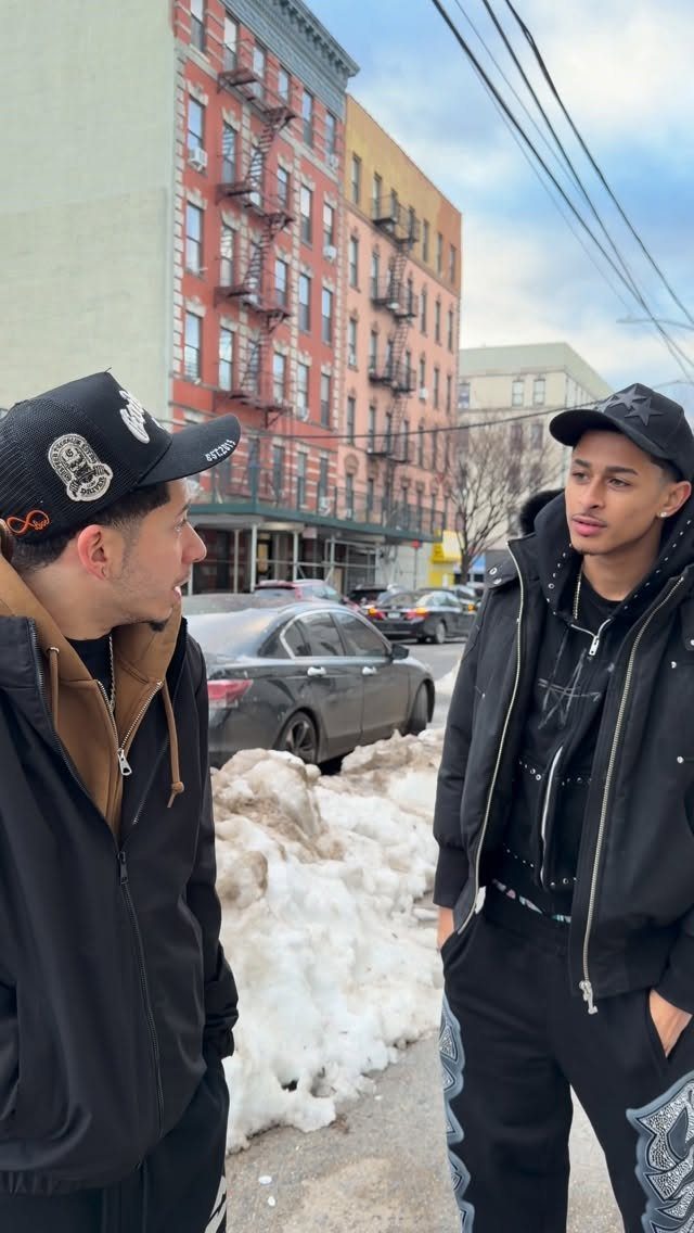 Two men in black winter streetwear standing on a snowy New York City street.