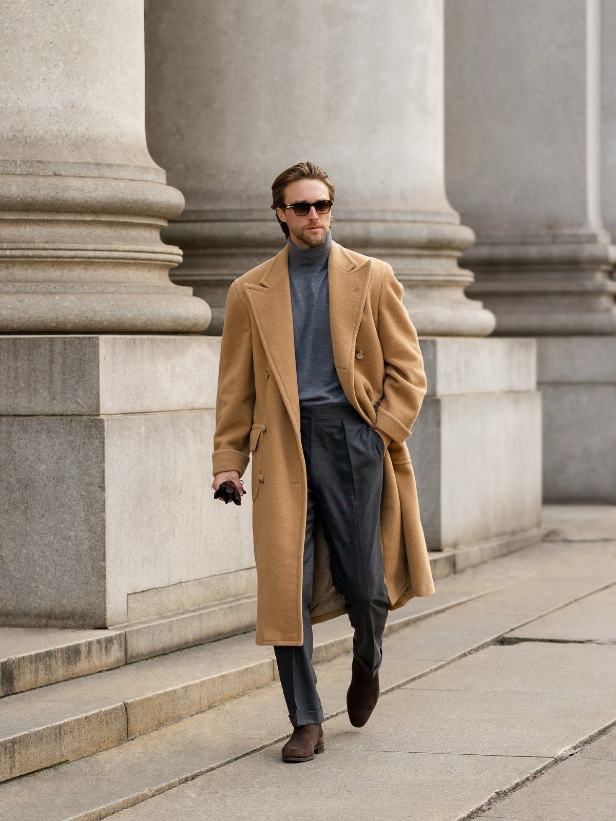 Man in a long tan wool coat and grey turtleneck walking past large stone columns.