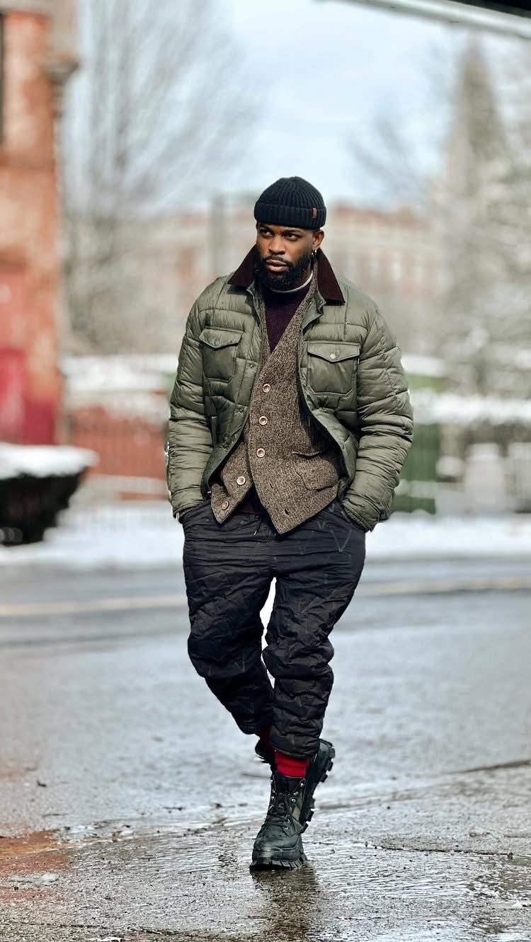 A man walking on a wet city street wearing a quilted olive jacket and black beanie.
