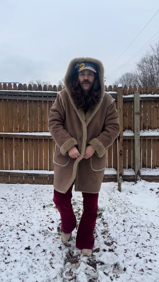 A man wearing a tan shearling coat and maroon trousers stands in a snowy yard.