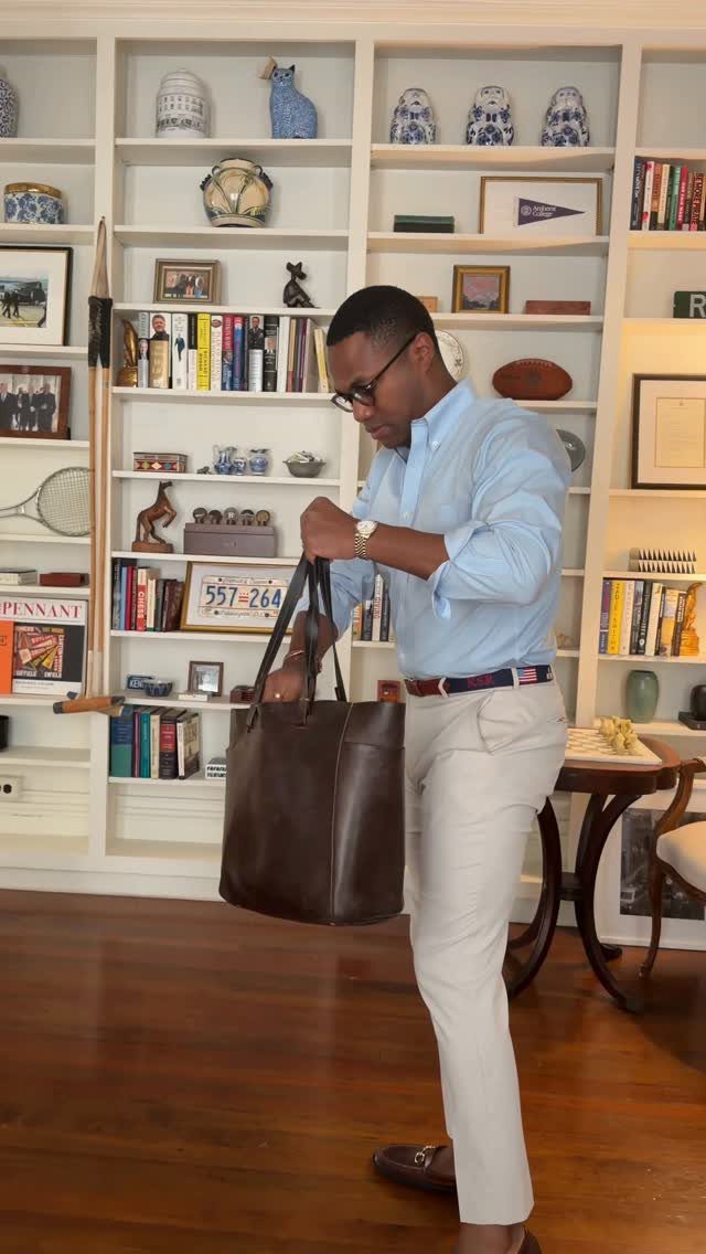 A man in a light blue shirt and beige chinos holds a large brown leather tote bag.