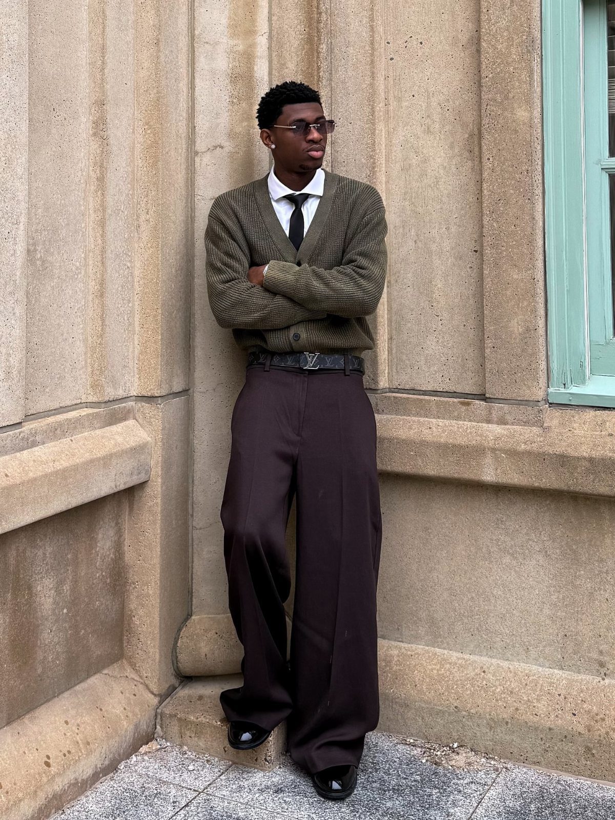 Man in an olive cardigan, white shirt, black tie, and wide brown trousers leaning against a stone wall.