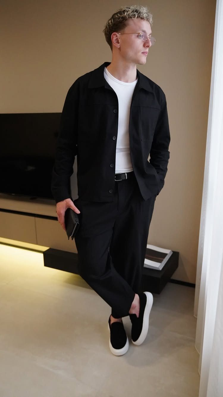 A young man wearing a black jacket, white t-shirt, black trousers, and black slip-on shoes.