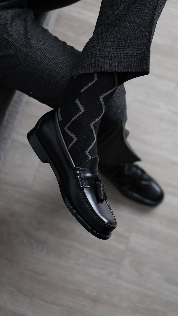 Black leather loafers worn with patterned socks and dark grey trousers on wood flooring.