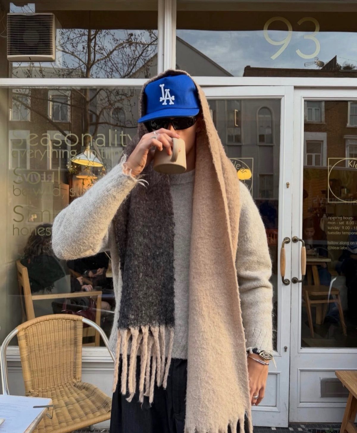 Person wearing a warm cream sweater and a large fringed scarf while holding a coffee cup.