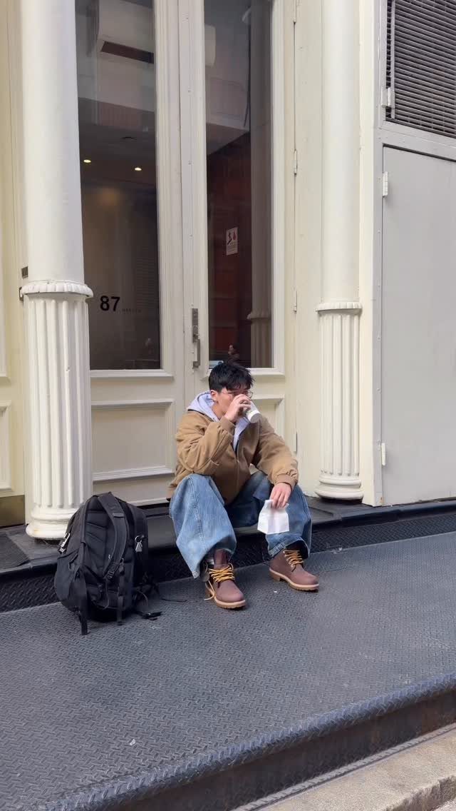 A person sitting on a doorstep wearing a tan jacket, hoodie, blue jeans, and brown boots.