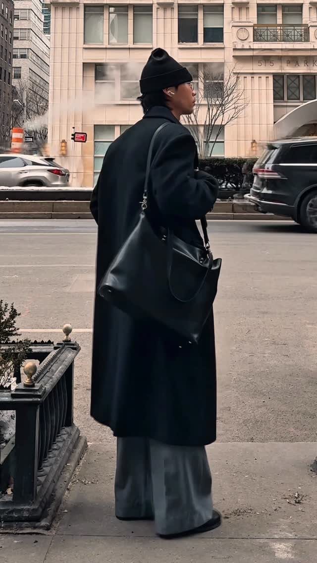 A person in a long black coat and beanie standing on a city street.