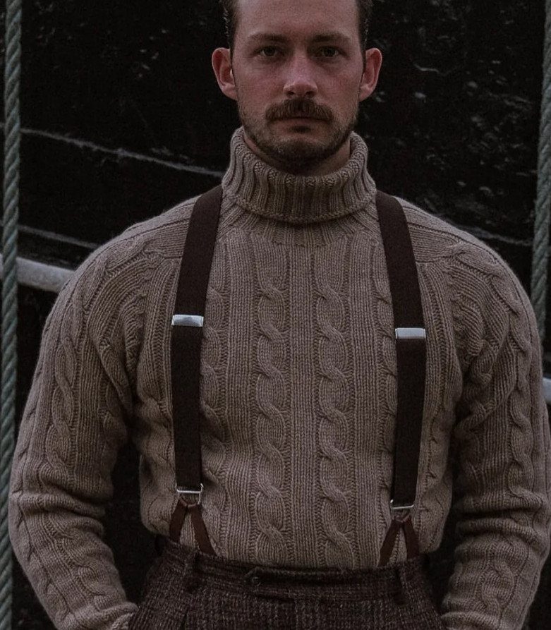 A man wearing a beige cable-knit wool turtleneck sweater and brown suspenders.