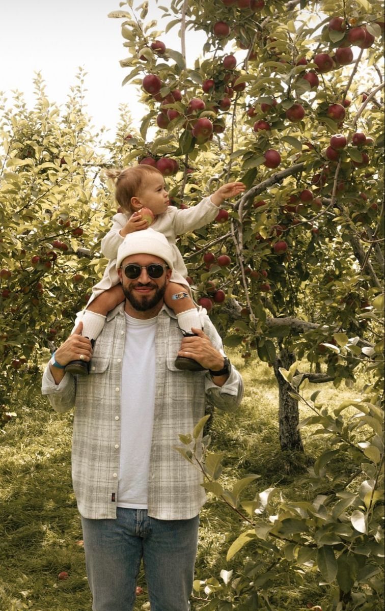 A man carrying a toddler on his shoulders while picking apples in an orchard.