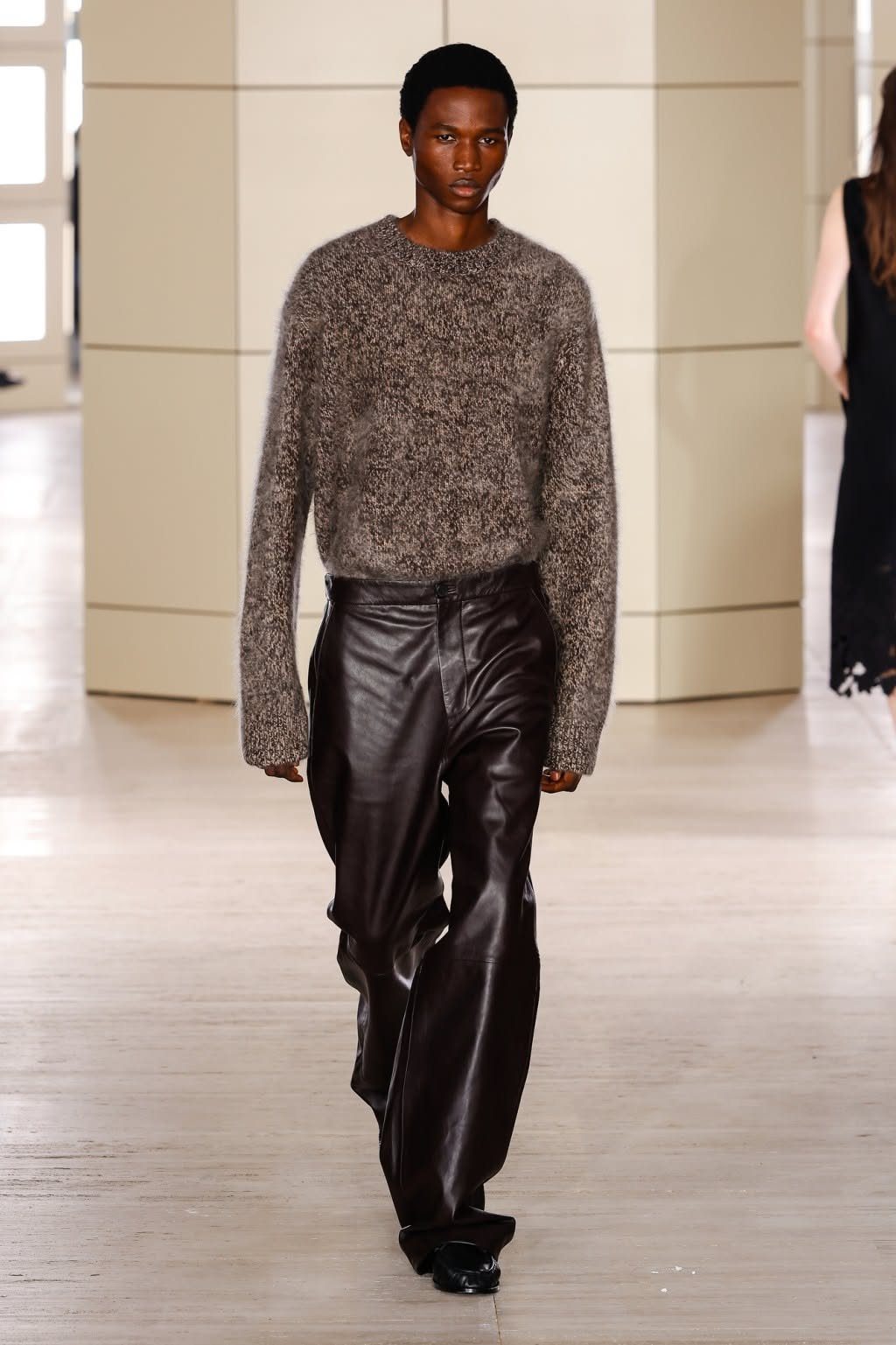A model walking a runway in a brown knit jumper and dark leather trousers.