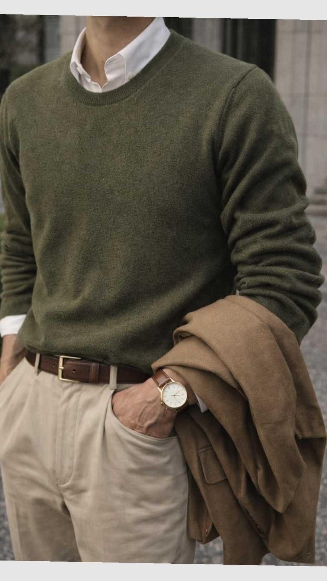Man in an olive green sweater and beige trousers holding a brown jacket.