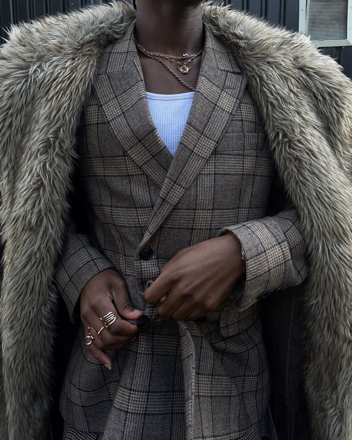 A person wearing a brown plaid blazer and a large faux-fur collar coat with gold jewelry.