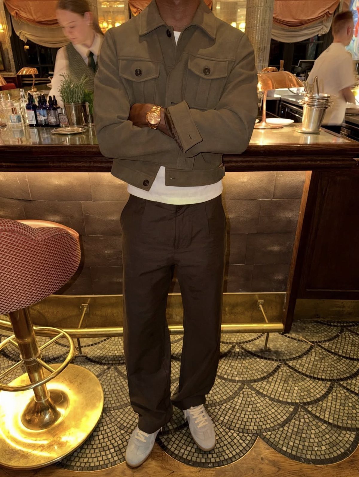 Man wearing an olive green jacket, dark trousers, and white sneakers standing in a bar.