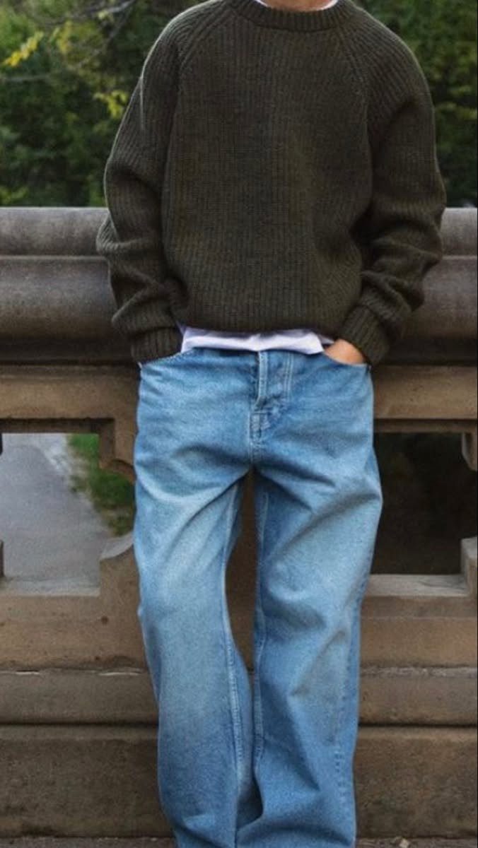 Person wearing an olive green ribbed sweater and wide-leg blue jeans standing by a stone wall.