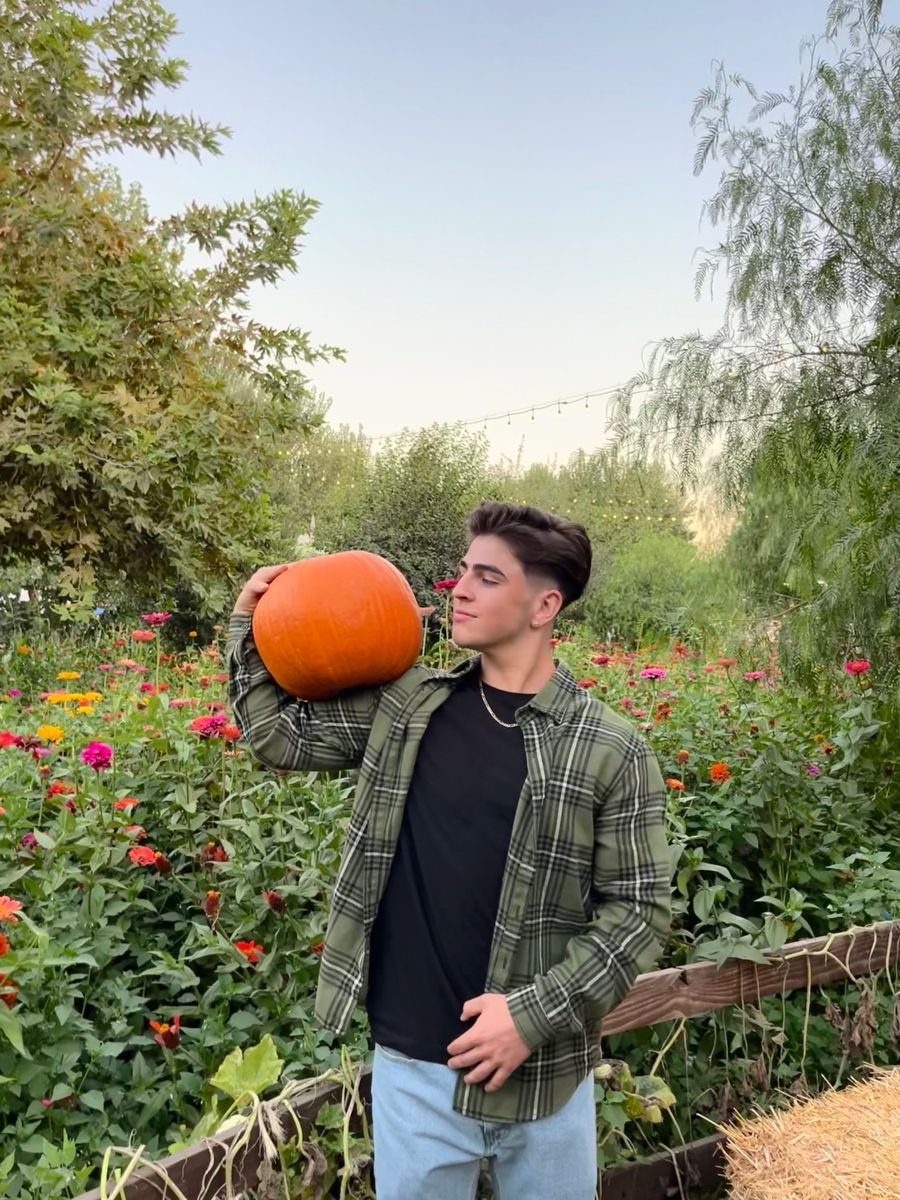 Young man holding a large orange pumpkin in a lush garden with colorful flowers.