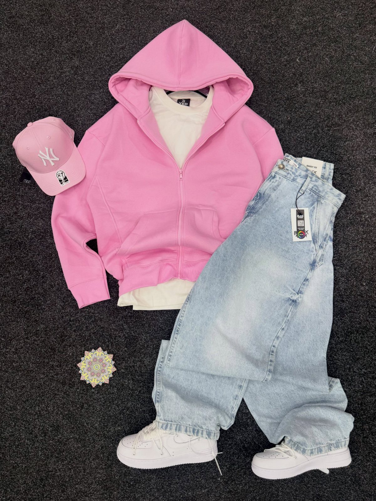 Pink zip-up hoodie, light blue baggy jeans, white sneakers, and a pink baseball cap laid on dark fabric.