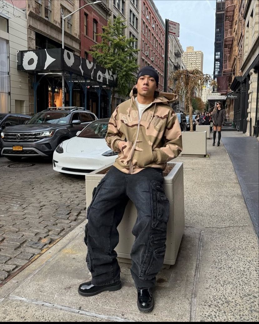 A man wearing an oversized tan hoodie and black cargo pants sitting on a city sidewalk.