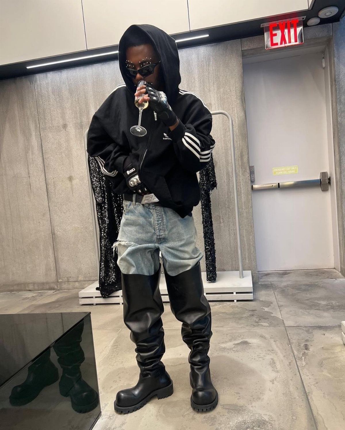 Person wearing a black hoodie, denim shorts, and black thigh-high boots holding a glass.