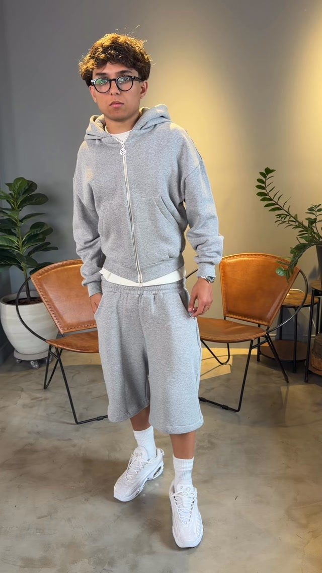 A young man wearing a grey oversized hoodie and matching shorts with white sneakers.