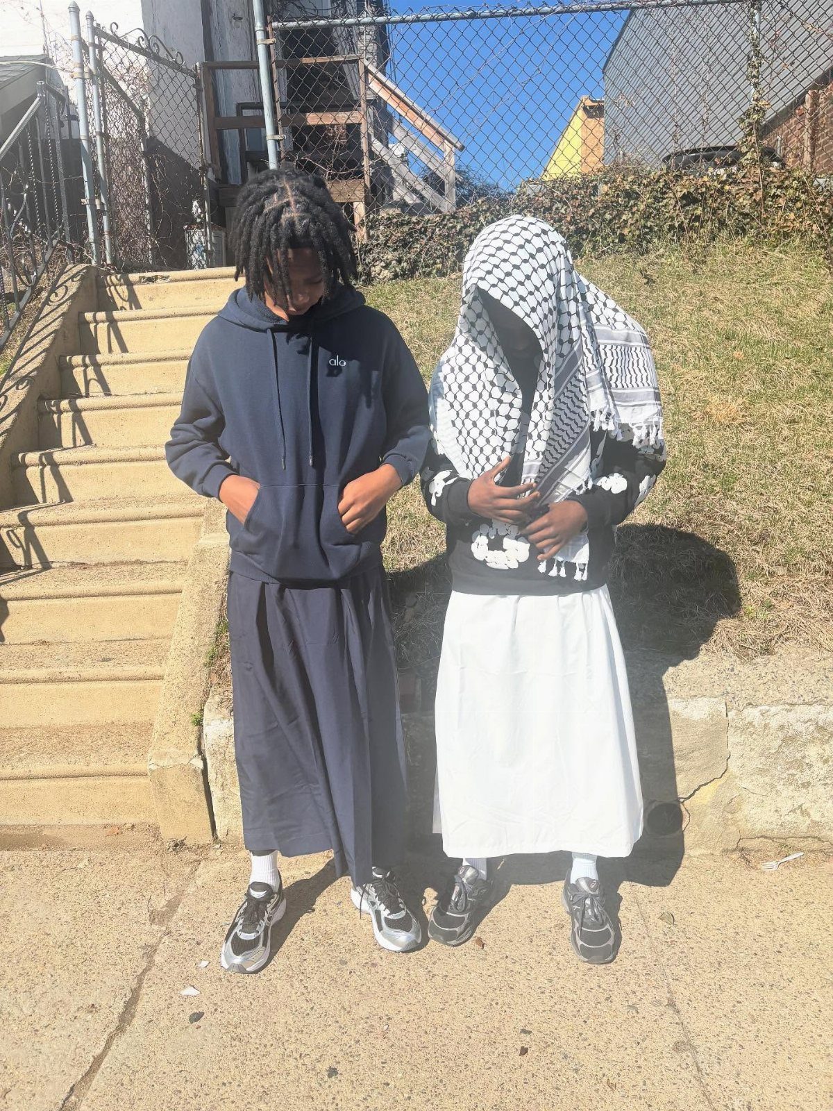 Two people standing outdoors wearing oversized hoodies and long skirts in bright sunlight.