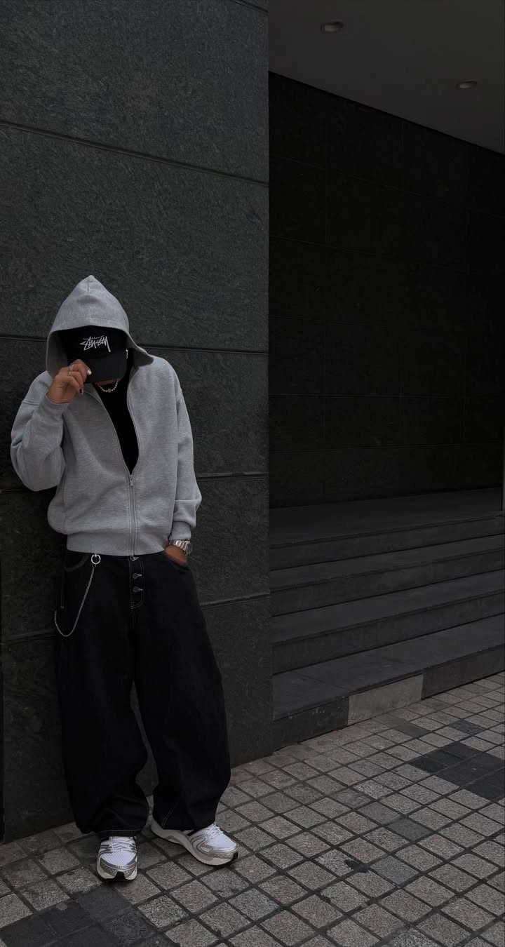 Person in a grey hoodie and black wide-leg pants leaning against a dark wall.
