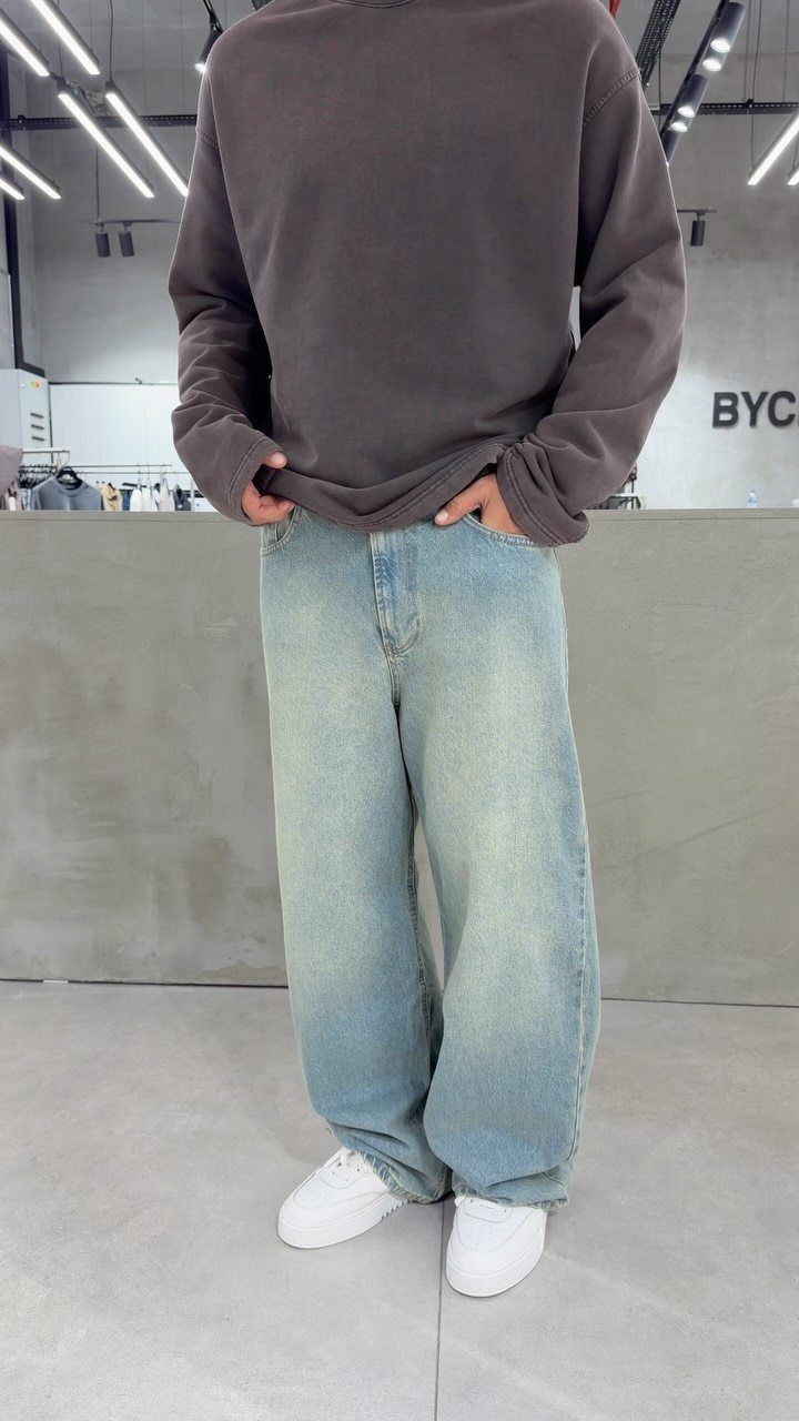 A person wearing a dark grey oversized sweatshirt, baggy light blue jeans, and white sneakers.