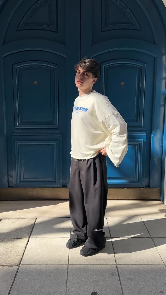 A young man posing in an oversized cream sweatshirt and loose black trousers against blue doors.