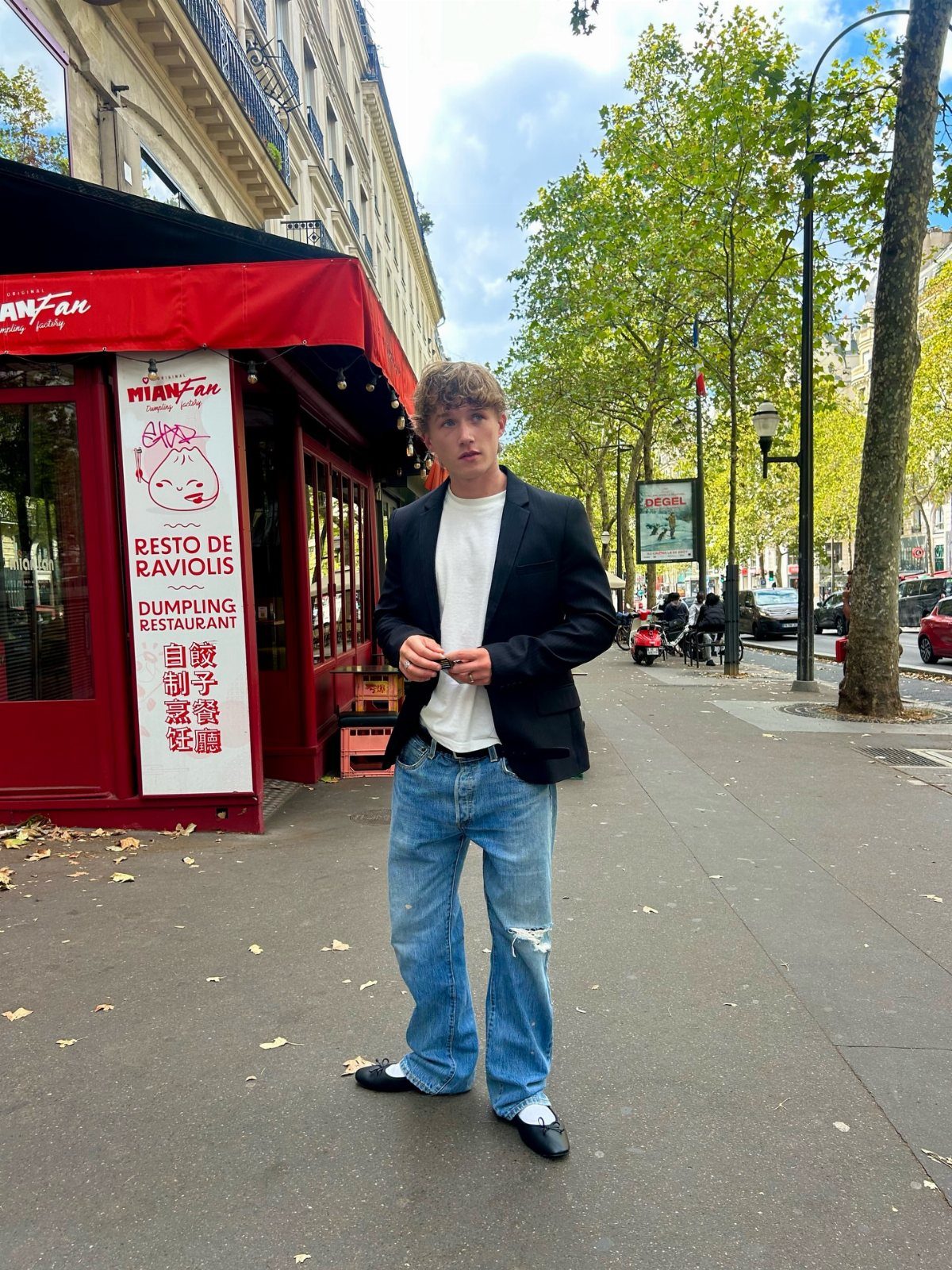 A man wearing a black blazer, blue jeans, and black ballet flats standing on a city sidewalk.
