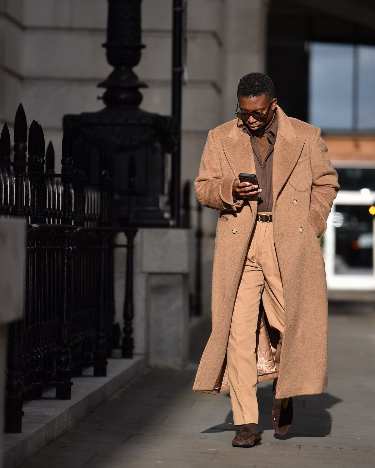 A man in a long tan coat and trousers walking while looking at his phone.