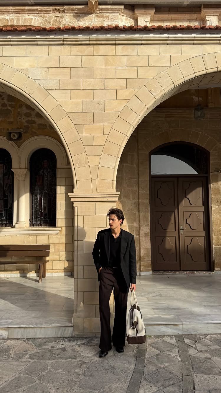 A person wearing a black oversized blazer and dark trousers standing by stone arches.