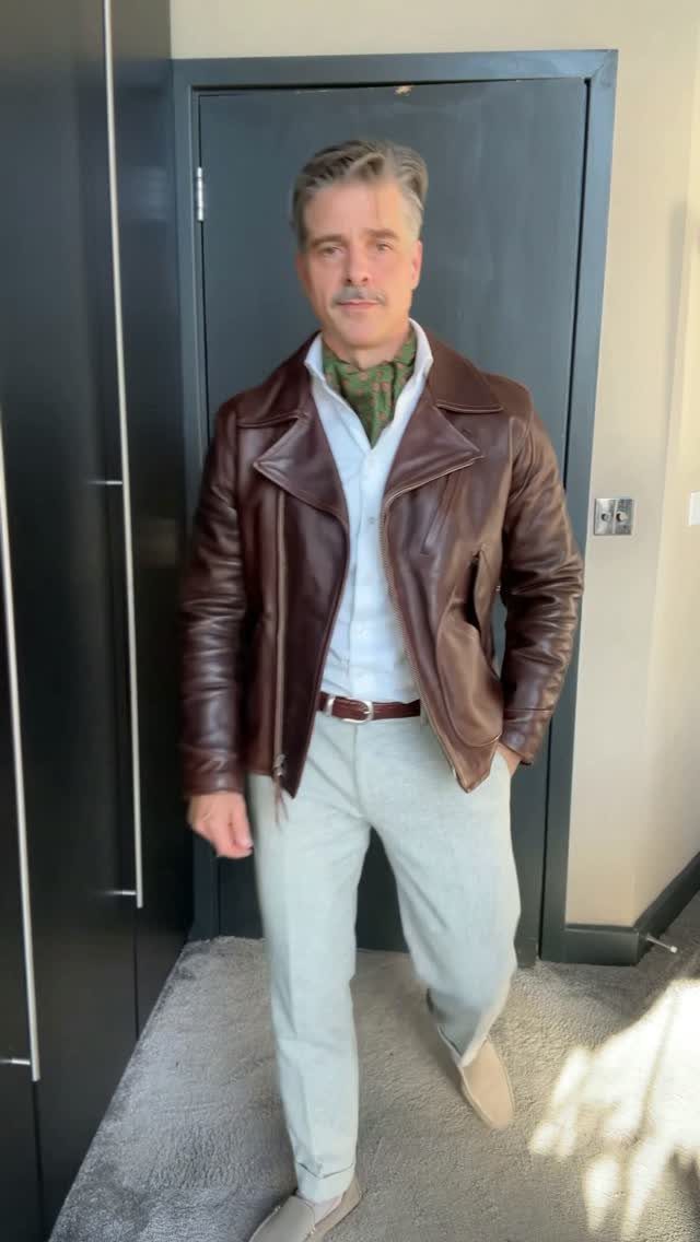 A man wearing a brown leather jacket and an olive green silk scarf around his neck.