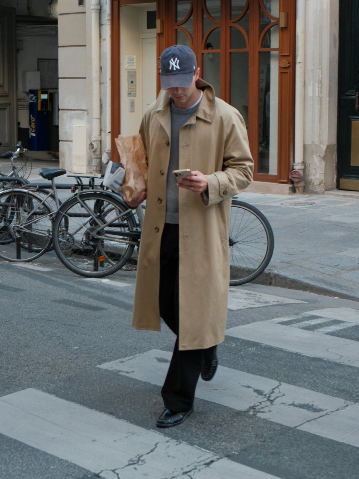 A man in a tan trench coat walking across a city street while using his phone.