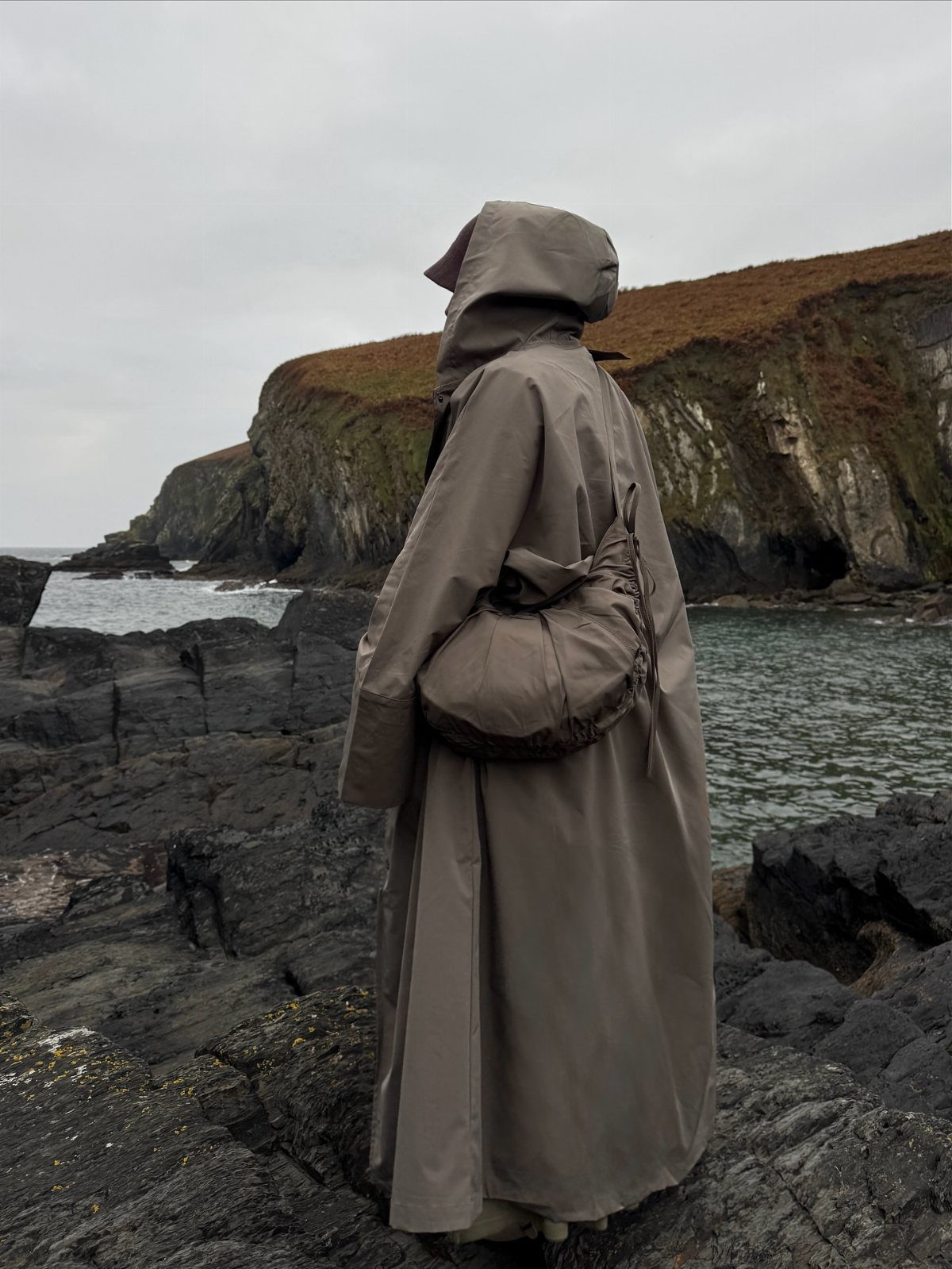 A person in a long hooded taupe raincoat standing on rocky coastal cliffs by the sea.