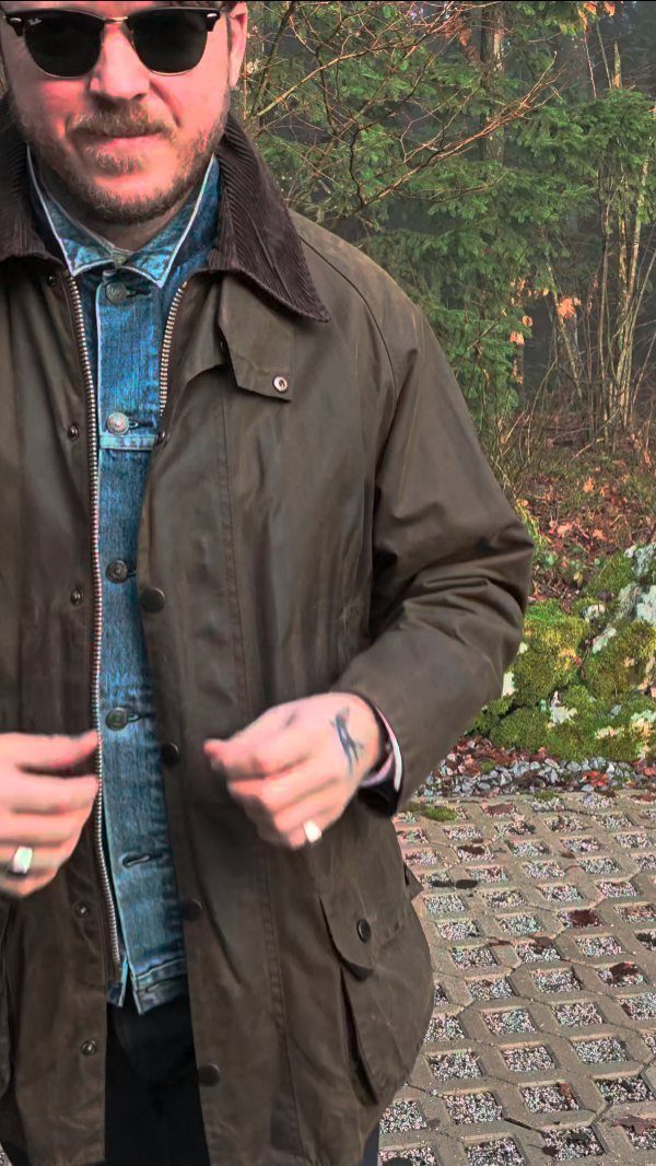A man wearing a dark brown hooded rain jacket and sunglasses standing in a garden.