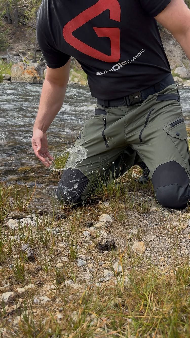 Man kneeling by a stream as water splashes against his olive green water-resistant cargo pants.